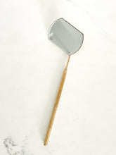 Load image into Gallery viewer, A large, rectangular lash mirror with a gold finish and an extendable golden handle, placed on a white surface.